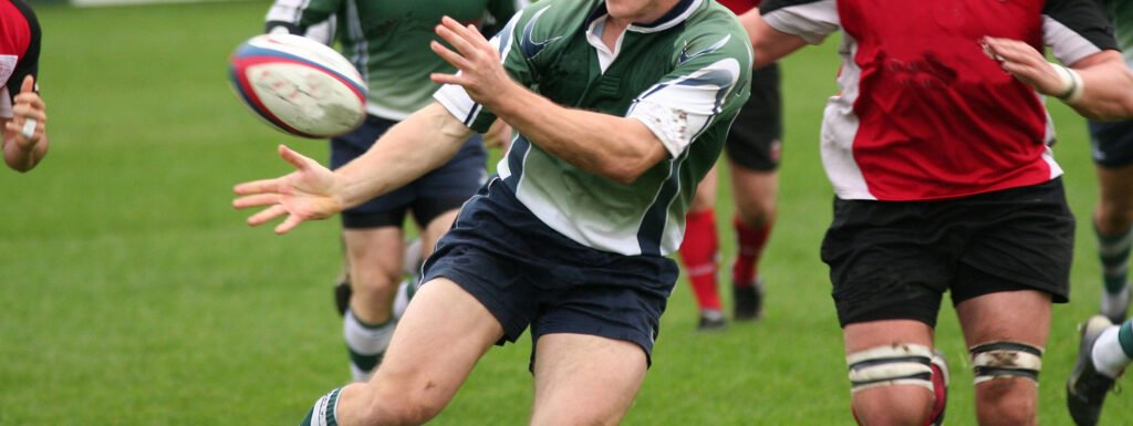 Photo of rugby players on field, centre player is catching the ball while running.