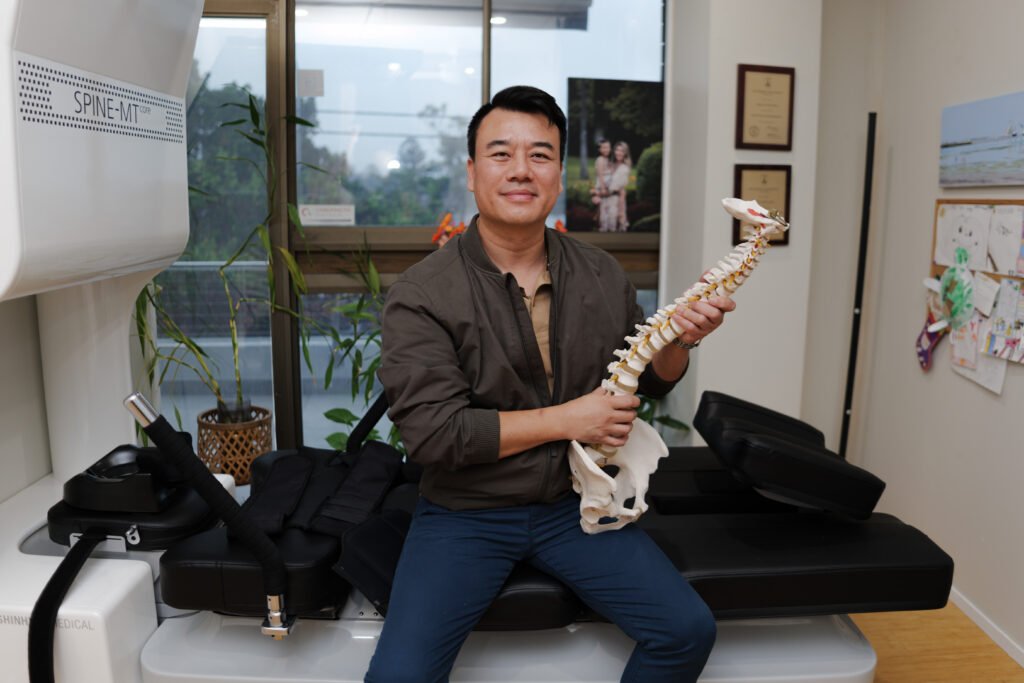 Taringa Chiropractor Brandon Ngo seated in his clinic, holding an anatomical spinal model.