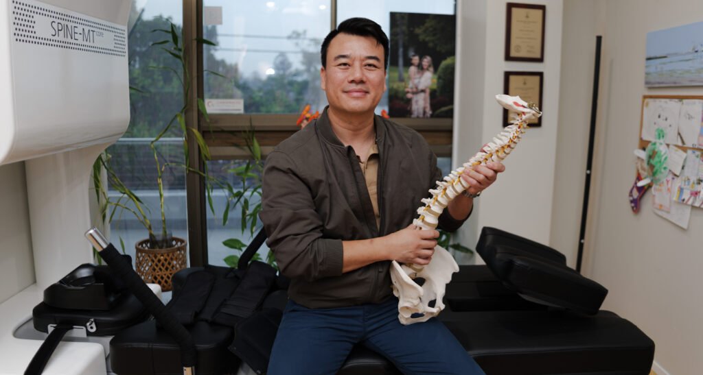 Chiropractor Brandon Ngo in his clinic, seated on the massage bench and smiling with a spinal anatomy model in his hands.